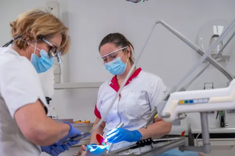 Tandarts hardt een witte vulling uit met de blauwe lamp, assistent houdt de tand droog, lichte behandelkamer