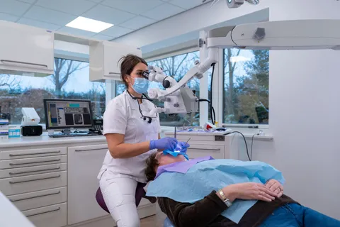 Tandarts werkt geconcentreerd met de behandelmicroscoop, assistent ernaast, moderne lichte behandelkamer