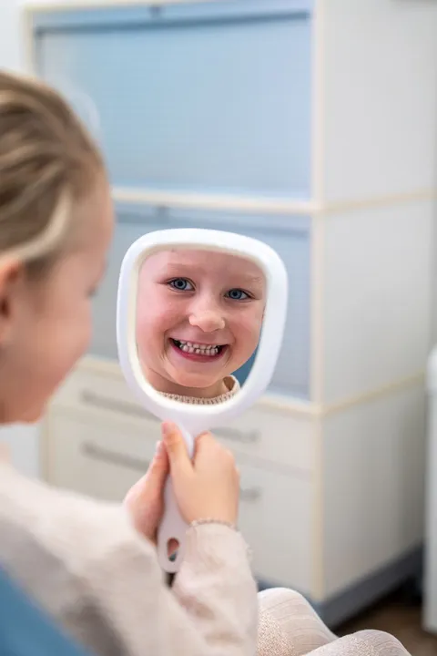 Kind in de stoel kijkt lachend mee in het spiegeltje, preventieassistent ernaast